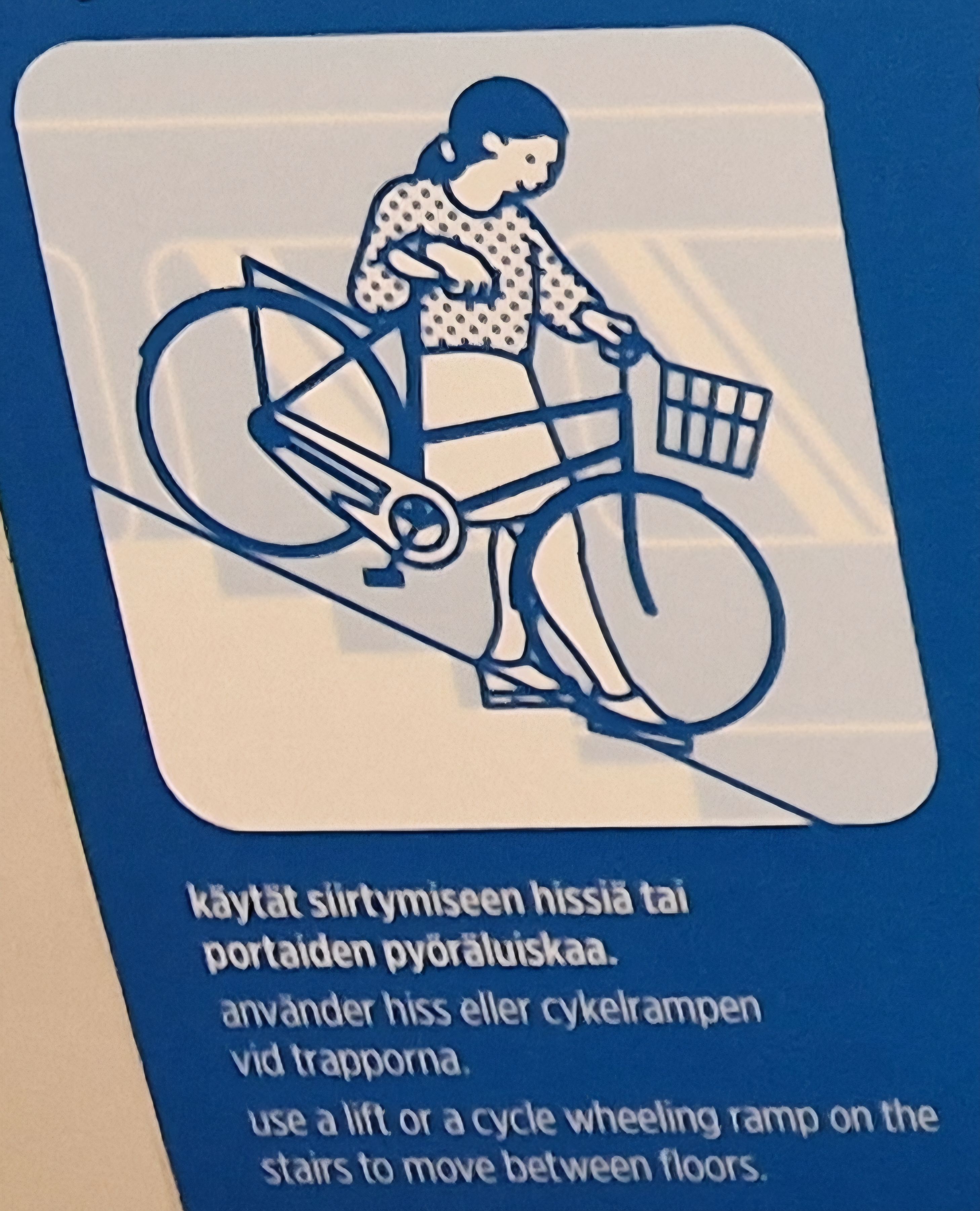 Hahmo taluttaa pyöräänsä pyöräluiskaa pitkin alaspäin portaissa. Alla lukee teksti 'käytät siirtymiseen hissiä tai portaiden pyöräluiskaa'.